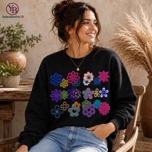 Puede incluir: Una sudadera negra de manga larga con una cuadrícula de flores bordadas de colores en tonos rosa, azul, morado, amarillo y blanco. El diseño consiste en varias formas florales dispuestas en un patrón rectangular en el pecho.