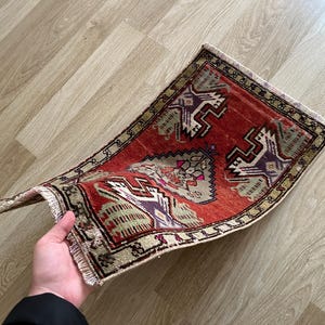 May include: A small, rectangular, hand-woven rug with a red background and intricate geometric patterns in beige, purple, and green. The rug has a fringed edge and is slightly curved, showcasing its texture and design.