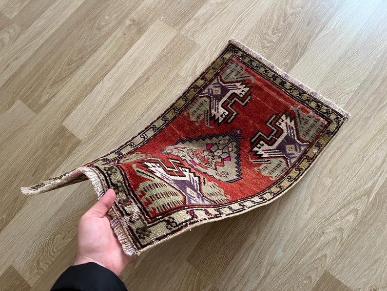 May include: A small, rectangular, hand-woven rug with a red background and intricate geometric patterns in beige, purple, and green. The rug has a fringed edge and is slightly curved, showcasing its texture and design.