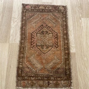 May include: A rectangular woven rug featuring a central geometric medallion in shades of brown, rust orange, and beige. The rug has a detailed border with repeating patterns and short fringe along the top and bottom edges.