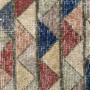 K&ouml;nnte beinhalten: Nahaufnahme eines Vintage-Teppichs mit geometrischem Muster. Der Teppich weist ein sich wiederholendes Design aus Dreiecken in Rot-, Blau-, Beige- und Gr&uuml;nt&ouml;nen auf. Der Teppich hat ein strukturiertes Aussehen mit sichtbaren gewebten F&auml;den.