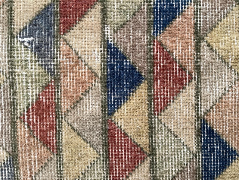 K&ouml;nnte beinhalten: Nahaufnahme eines Vintage-Teppichs mit geometrischem Muster. Der Teppich weist ein sich wiederholendes Design aus Dreiecken in Rot-, Blau-, Beige- und Gr&uuml;nt&ouml;nen auf. Der Teppich hat ein strukturiertes Aussehen mit sichtbaren gewebten F&auml;den.