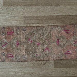 May include: A rectangular, vintage-style rug with a faded, distressed appearance. The rug features a muted color palette of pink, gray, and brown, with geometric and floral patterns in pink. The edges have a fringed detail.