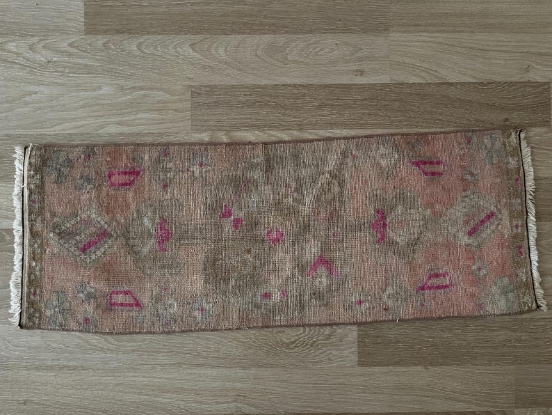 May include: A rectangular, vintage-style rug with a faded, distressed appearance. The rug features a muted color palette of pink, gray, and brown, with geometric and floral patterns in pink. The edges have a fringed detail.