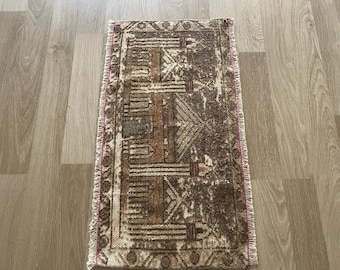 Op de afbeelding: Een rechthoekig, versleten tapijt met een geometrisch patroon in bruin-, beige- en roze tinten. Het tapijt heeft een gefranjerde rand en ligt op een lichtgekleurde houten vloer. Het ontwerp toont lijnen en vormen.