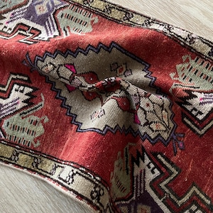 May include: A distressed, vintage-style rug with a rich red field and intricate geometric patterns in beige, purple, and green. The rug features a central medallion design and a detailed border, showcasing a traditional aesthetic.