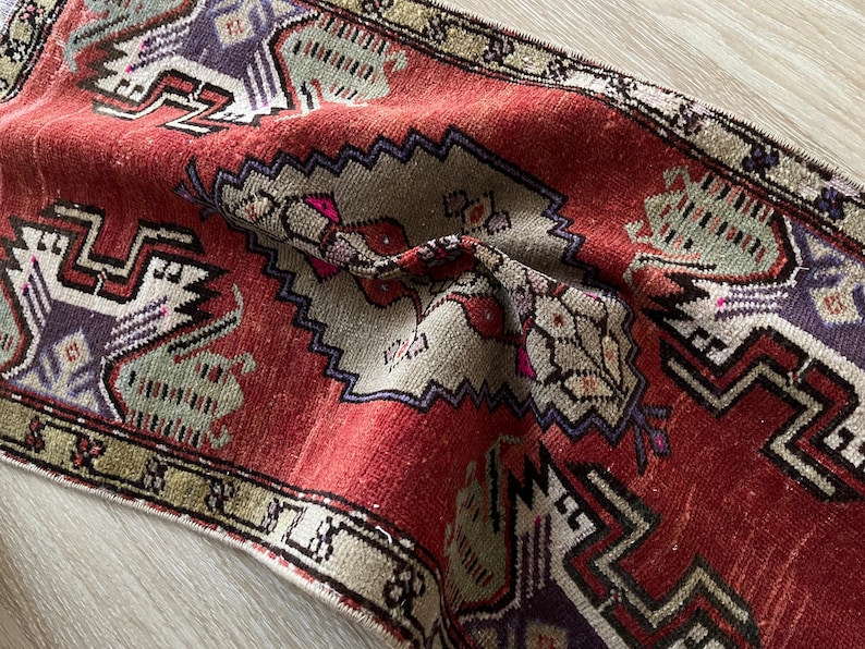 May include: A distressed, vintage-style rug with a rich red field and intricate geometric patterns in beige, purple, and green. The rug features a central medallion design and a detailed border, showcasing a traditional aesthetic.