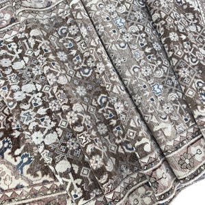 May include: A woven area rug featuring a complex geometric and floral pattern in shades of brown, beige, cream, and muted blue. The design includes repeating diamond shapes, intricate borders, and stylized botanical motifs across the surface.