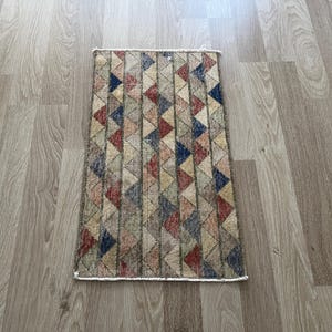 K&ouml;nnte beinhalten: Rechteckiger Teppich mit einem geometrischen Muster aus Dreiecken in Rot-, Blau-, Beige- und Gr&uuml;nt&ouml;nen. Der Teppich hat einen cremefarbenen Rand und ein verwittertes, Vintage-Aussehen. Der Teppich liegt auf einem hellen Holzboden.