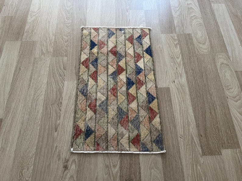 K&ouml;nnte beinhalten: Rechteckiger Teppich mit einem geometrischen Muster aus Dreiecken in Rot-, Blau-, Beige- und Gr&uuml;nt&ouml;nen. Der Teppich hat einen cremefarbenen Rand und ein verwittertes, Vintage-Aussehen. Der Teppich liegt auf einem hellen Holzboden.