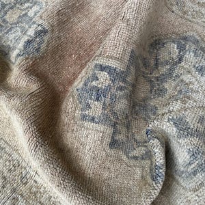 Peut inclure: Un tapis tissé présentant une couleur de base beige et brun clair délavée avec des motifs floraux et géométriques complexes et usés dans des tons bleu ardoise sourds. Le textile a une texture veloutée visible et est plié.