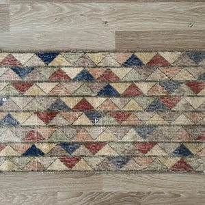 K&ouml;nnte beinhalten: Rechteckiger Teppich mit einem geometrischen Muster aus roten, blauen, beigen und gr&uuml;nen Dreiecken. Der Teppich hat einen Used-Look mit wei&szlig;em Fransenbesatz. Er liegt auf einem hellen Holzboden.