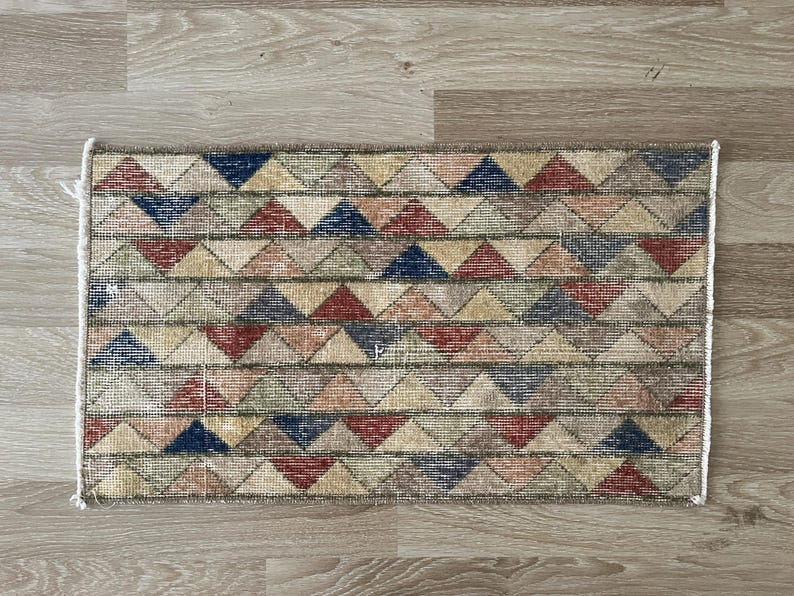 K&ouml;nnte beinhalten: Rechteckiger Teppich mit einem geometrischen Muster aus roten, blauen, beigen und gr&uuml;nen Dreiecken. Der Teppich hat einen Used-Look mit wei&szlig;em Fransenbesatz. Er liegt auf einem hellen Holzboden.
