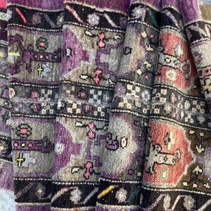 May include: A woven textile rug featuring a repeating pattern of geometric shapes and floral motifs in shades of deep purple, charcoal gray, muted terracotta, and black. The fabric has a textured, ribbed surface with intricate, symmetrical designs.