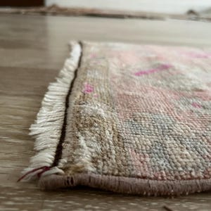 May include: Close-up of a small, rectangular rug with a distressed, vintage appearance. The rug features a beige background with pink and blue accents. The edges are finished with a brown border and white fringe.