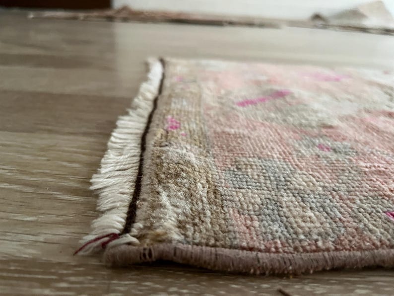 May include: Close-up of a small, rectangular rug with a distressed, vintage appearance. The rug features a beige background with pink and blue accents. The edges are finished with a brown border and white fringe.