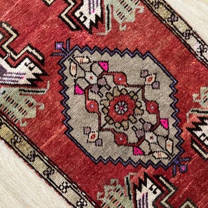 May include: A close-up of a red and gray rug with a central geometric design. The rug features a central medallion with a floral pattern in shades of pink, red, and gray. The border is a darker red with geometric shapes.