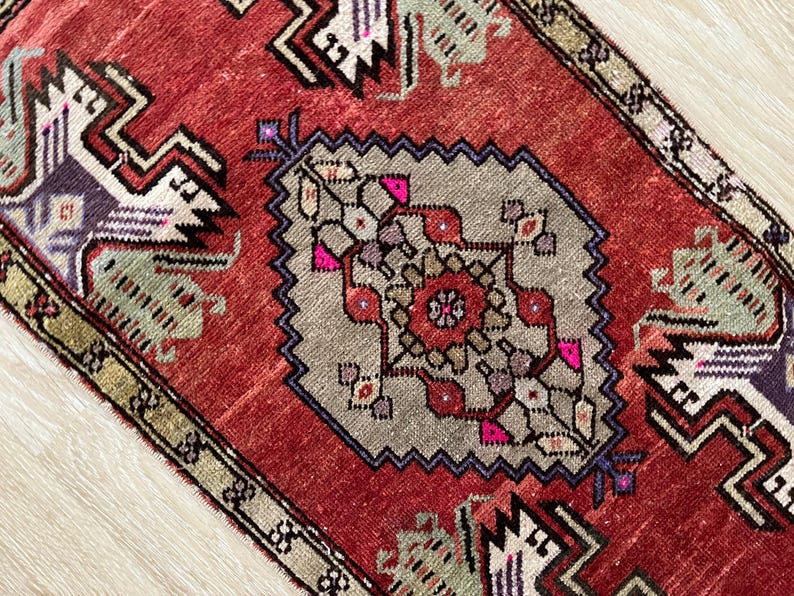 May include: A close-up of a red and gray rug with a central geometric design. The rug features a central medallion with a floral pattern in shades of pink, red, and gray. The border is a darker red with geometric shapes.