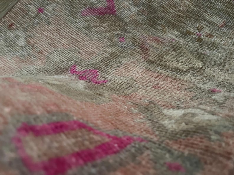 May include: Close-up of a woven textile with a textured surface. The fabric features a blend of muted colors, including gray, pink, and beige, with accents of bright fuchsia. The design appears to be a rug or textile.