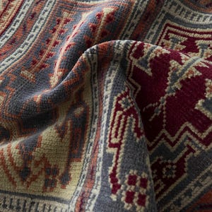 May include: A woven rug featuring a dense pile with a complex geometric pattern in deep red, muted blue-grey, and soft beige. The fabric is folded to reveal the textured surface, showcasing intricate repeating motifs and linear border designs.
