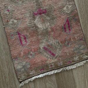 May include: A small, rectangular rug with a faded, vintage appearance. The rug features a blend of pink, gray, and brown hues, with accents of fuchsia. The design includes geometric shapes and a fringed edge, suggesting a traditional style.