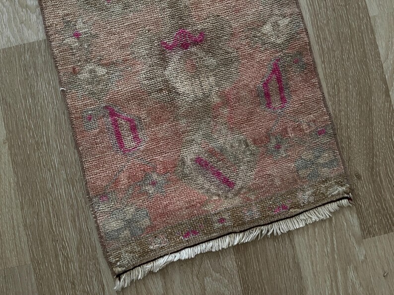May include: A small, rectangular rug with a faded, vintage appearance. The rug features a blend of pink, gray, and brown hues, with accents of fuchsia. The design includes geometric shapes and a fringed edge, suggesting a traditional style.