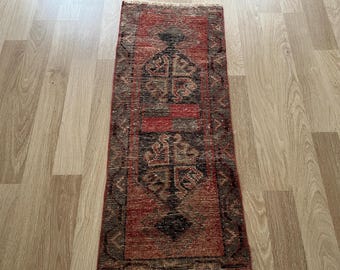 Puede incluir: Una alfombra rectangular vintage con aspecto desgastado. La alfombra presenta un diseño central en tonos rojos, negros y marrones, con intrincados patrones geométricos. La alfombra tiene un borde con flecos y está colocada sobre un suelo de madera clara.
