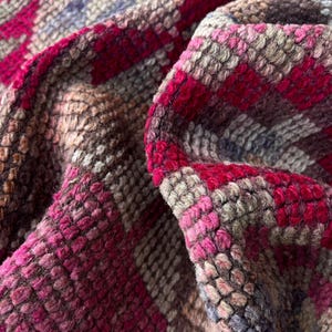 May include: A thick, textured textile featuring a repeating geometric pattern in shades of vibrant magenta, deep pink, muted brown, and soft gray. The fabric has a dense, looped pile construction with visible folds that create depth and shadows.