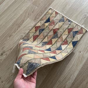 K&ouml;nnte beinhalten: Ein kleiner, rechteckiger Teppich mit einem geometrischen Muster aus Dreiecken in Rot-, Blau-, Gelb- und Beiget&ouml;nen. Der Teppich hat einen Fransenrand und scheint handgefertigt zu sein.