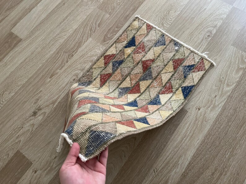 K&ouml;nnte beinhalten: Ein kleiner, rechteckiger Teppich mit einem geometrischen Muster aus Dreiecken in Rot-, Blau-, Gelb- und Beiget&ouml;nen. Der Teppich hat einen Fransenrand und scheint handgefertigt zu sein.