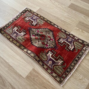 May include: A rectangular, hand-woven rug with a red background and intricate geometric patterns in shades of purple, beige, and green. The rug has a fringed edge and measures approximately 24 x 36 inches.
