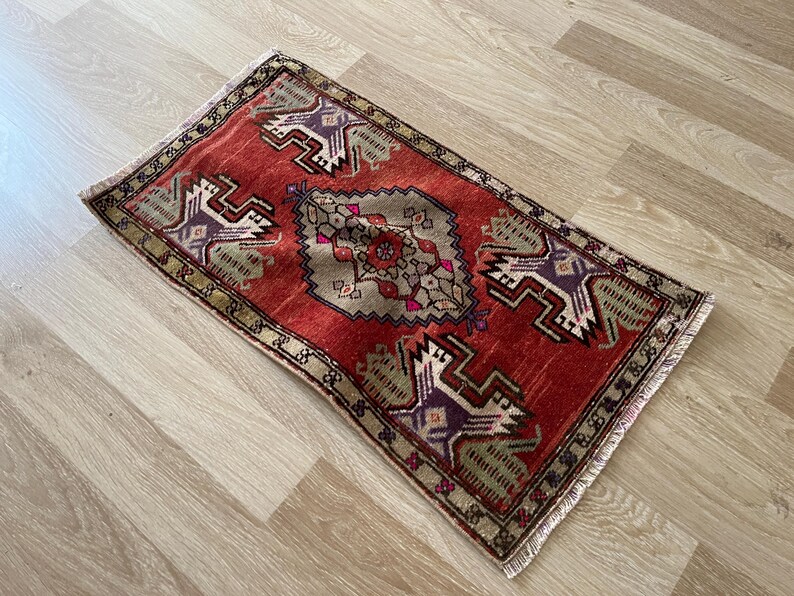 May include: A rectangular, hand-woven rug with a red background and intricate geometric patterns in shades of purple, beige, and green. The rug has a fringed edge and measures approximately 24 x 36 inches.