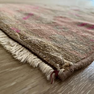 May include: Close-up of a rectangular rug with a distressed, vintage appearance. The rug features a mix of muted colors, including beige, brown, and pink, with a fringed edge. The rug is placed on a light-colored wooden surface.