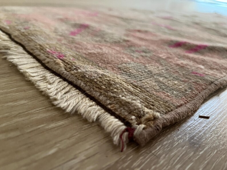 May include: Close-up of a rectangular rug with a distressed, vintage appearance. The rug features a mix of muted colors, including beige, brown, and pink, with a fringed edge. The rug is placed on a light-colored wooden surface.