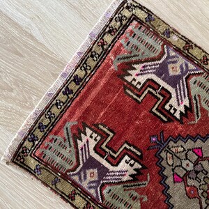 May include: A small, rectangular rug with a detailed geometric pattern. The rug features a red background with accents of purple, gray, and green. The border is a mix of green and gold, with a fringed edge.