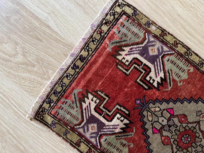 May include: A small, rectangular rug with a detailed geometric pattern. The rug features a red background with accents of purple, gray, and green. The border is a mix of green and gold, with a fringed edge.