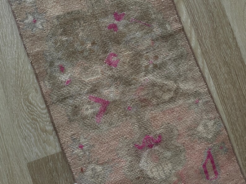 May include: A rectangular, vintage-style rug with a faded, distressed appearance. The rug features a blend of muted colors, including pink, brown, and gray, with accents of brighter pink floral patterns. The rug's texture appears soft and worn.
