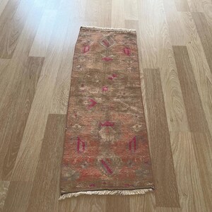 May include: A rectangular, distressed-look rug with a brown and reddish-orange color scheme. The rug features a repeating pattern of geometric shapes and floral motifs in pink and green. The rug has a fringed edge.