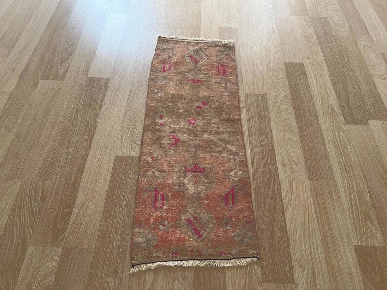 May include: A rectangular, distressed-look rug with a brown and reddish-orange color scheme. The rug features a repeating pattern of geometric shapes and floral motifs in pink and green. The rug has a fringed edge.