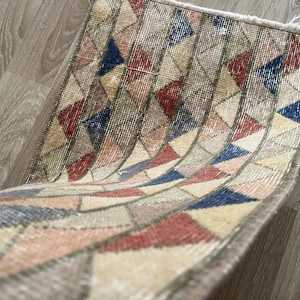 K&ouml;nnte beinhalten: Kleiner, rechteckiger Teppich mit einem geometrischen Muster aus roten, blauen, beigen und grauen Dreiecken. Der Teppich hat einen Used-Look im Vintage-Stil mit ausgefransten Kanten. Der Teppich ist gefaltet und zeigt seine Unterseite.