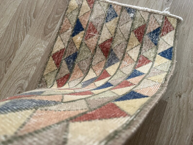 K&ouml;nnte beinhalten: Kleiner, rechteckiger Teppich mit einem geometrischen Muster aus roten, blauen, beigen und grauen Dreiecken. Der Teppich hat einen Used-Look im Vintage-Stil mit ausgefransten Kanten. Der Teppich ist gefaltet und zeigt seine Unterseite.