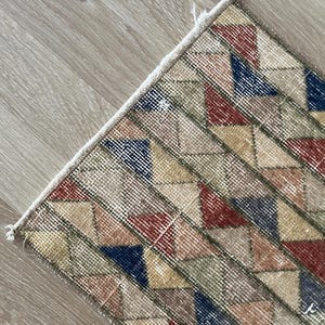 K&ouml;nnte beinhalten: Nahaufnahme eines Teils eines geometrisch gemusterten Teppichs. Der Teppich zeigt ein sich wiederholendes Muster aus Rauten in Rot-, Blau-, Gelb- und Gr&uuml;nt&ouml;nen auf beigem Hintergrund. Der Teppich hat eine strukturierte Oberfl&auml;che.