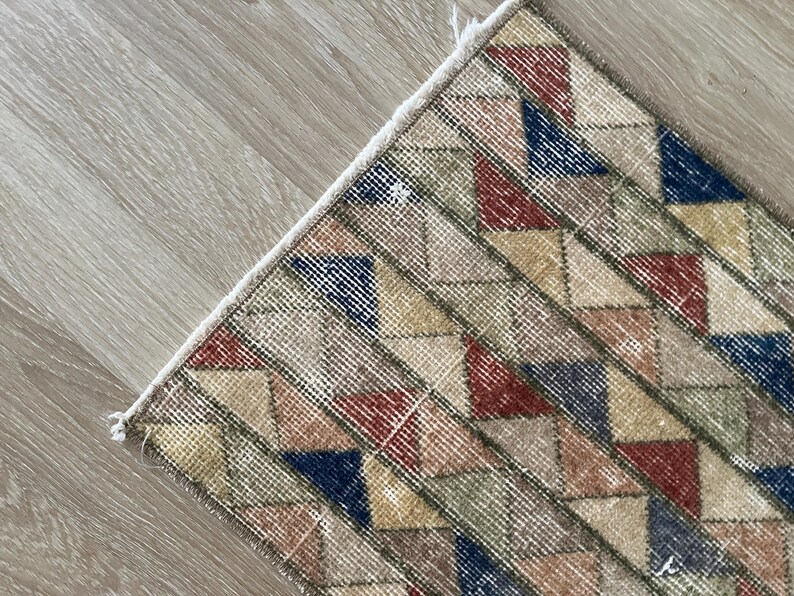 K&ouml;nnte beinhalten: Nahaufnahme eines Teils eines geometrisch gemusterten Teppichs. Der Teppich zeigt ein sich wiederholendes Muster aus Rauten in Rot-, Blau-, Gelb- und Gr&uuml;nt&ouml;nen auf beigem Hintergrund. Der Teppich hat eine strukturierte Oberfl&auml;che.