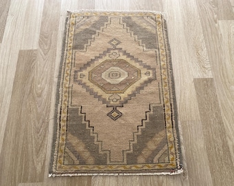 Puede incluir: Una alfombra tejida rectangular con un patrón central de medallón de diamante en tonos beige, topo y amarillo apagado. La alfombra presenta un borde geométrico y flecos cortos a lo largo de los bordes superior e inferior del diseño.