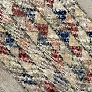 K&ouml;nnte beinhalten: Nahaufnahme eines rechteckigen Teppichs mit geometrischem Muster. Der Teppich weist ein sich wiederholendes Design aus Dreiecken in verschiedenen Farben auf, darunter Rot, Blau, Beige und Gr&uuml;n, vor einem neutralen Hintergrund. Der Teppich hat ein abgenutztes, Vintage-Aussehen.