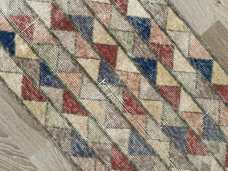 K&ouml;nnte beinhalten: Nahaufnahme eines rechteckigen Teppichs mit geometrischem Muster. Der Teppich weist ein sich wiederholendes Design aus Dreiecken in verschiedenen Farben auf, darunter Rot, Blau, Beige und Gr&uuml;n, vor einem neutralen Hintergrund. Der Teppich hat ein abgenutztes, Vintage-Aussehen.