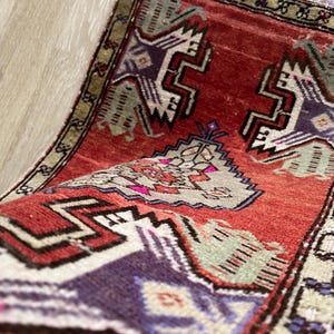 May include: A rolled-up, small, rectangular rug with a red background and geometric patterns in purple, white, and green. The rug has a beige border and a fringed edge. The design features stylized shapes.