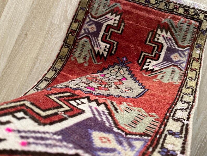 May include: A rolled-up, small, rectangular rug with a red background and geometric patterns in purple, white, and green. The rug has a beige border and a fringed edge. The design features stylized shapes.