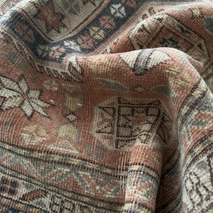 Può includere: Primo piano di un tappeto vintage con un motivo dettagliato e sbiadito. Il tappeto presenta un mix di colori caldi, tra cui ruggine, beige e blu tenui. Sono visibili intricati motivi geometrici e floreali.