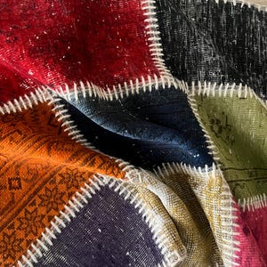 Op de afbeelding: Close-up van een patchwork tapijt met een verscheidenheid aan kleuren en patronen. Het tapijt heeft vierkanten van rood, oranje, zwart, blauw, groen en paars, aan elkaar gestikt met witte draad. Het tapijt heeft een vintage, distressed look.